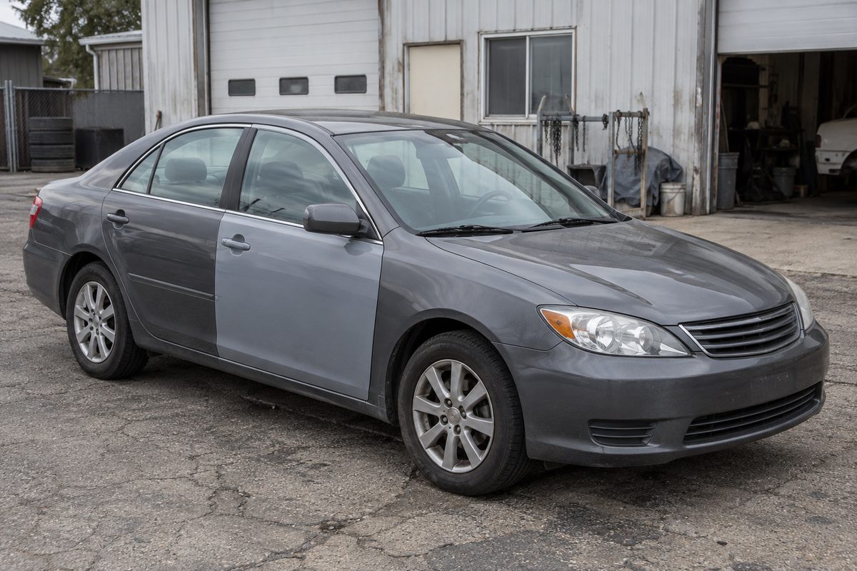 An older gray sedan with one lighter gray replacement front door, parked on cracked asphalt outside a metal-sided garage