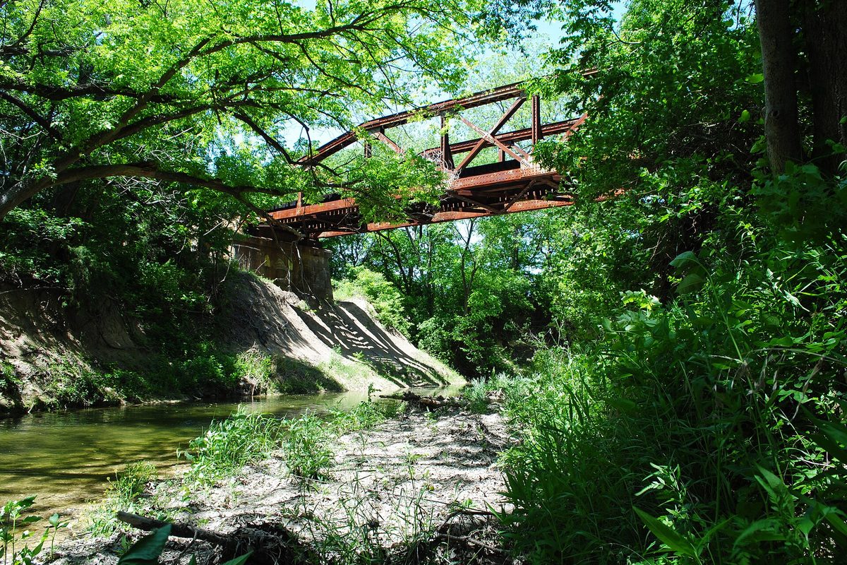 A rusty pony truss railroad bridge over Red Oak Creek, seen from the shaded creek bed with green trees and tall grass on both banks