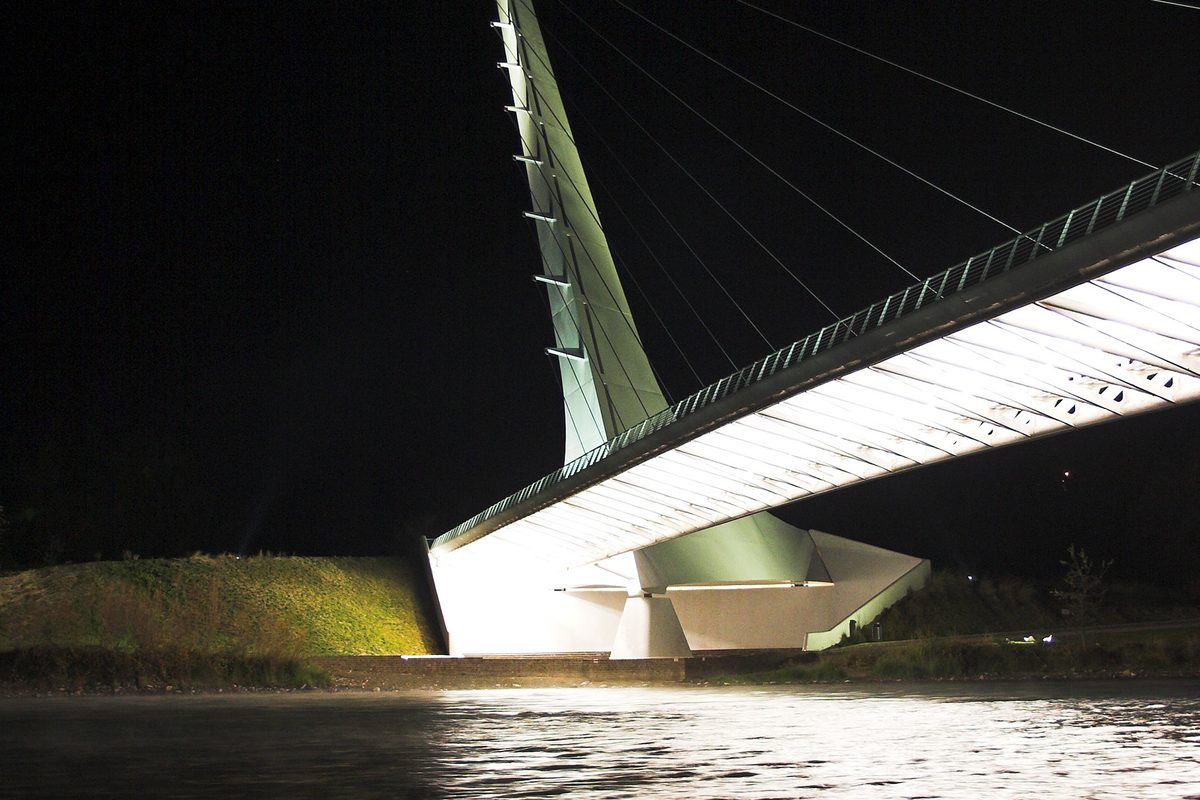 The Sundial Bridge in Redding lit up at night, its white angled pylon and cable-hung deck glowing above dark water