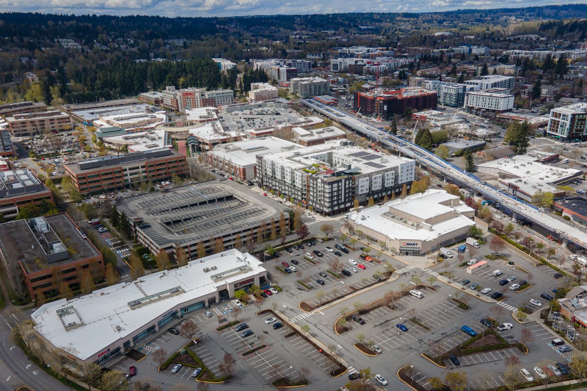 Drone view over downtown Redmond, with parking garages, apartment blocks and an elevated rail line curving between them, wooded hills beyond