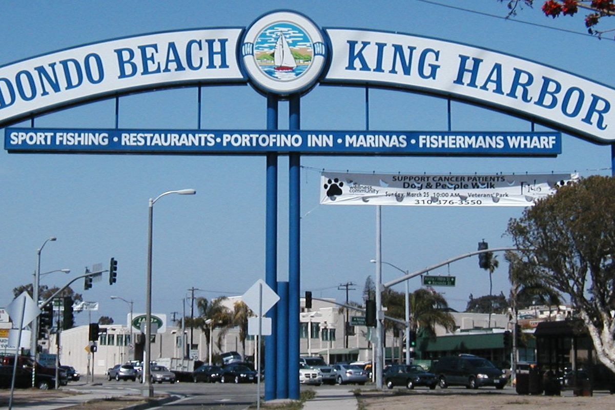 A blue arch sign reading Redondo Beach King Harbor, listing sport fishing, restaurants, marinas and Fishermans Wharf, over a street with parked cars