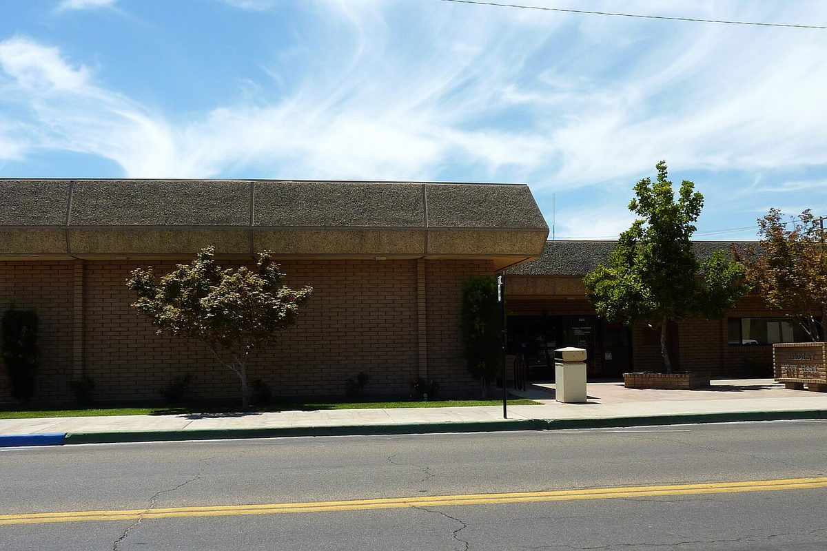 Reedley City Hall, a low brown brick building with a dark sloped roof, small trees in front and an empty two-lane street with a yellow center line