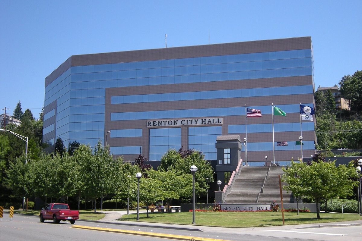 Renton City Hall, a tall building wrapped in bands of blue glass with its name over the entrance stairs, flags out front and a red pickup passing on the street