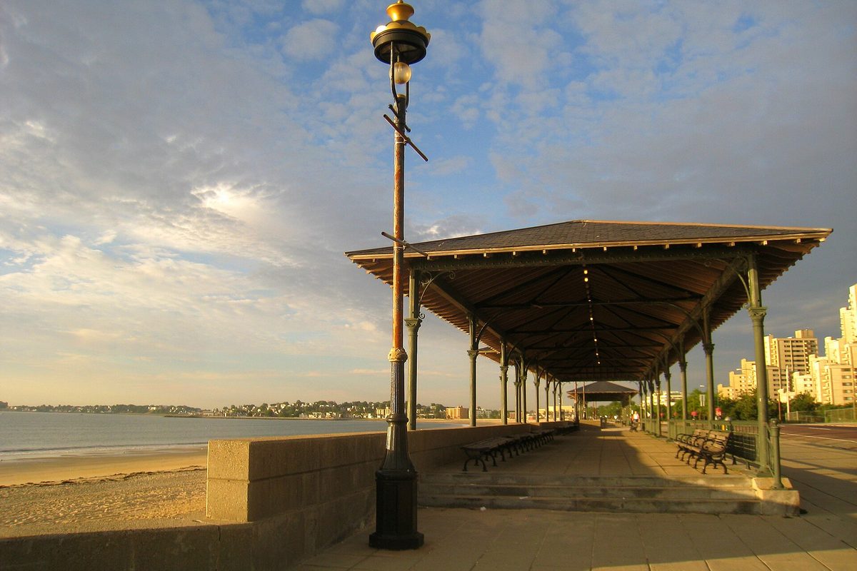 A covered pavilion with green iron posts on the Revere Beach boardwalk at sunrise, sand and calm water to the left and high-rises in the distance