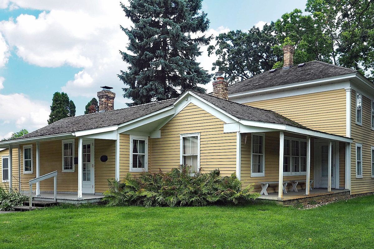 The yellow clapboard Riley Lucas Bartholomew House in Richfield, with white trim, two brick chimneys and a tall spruce behind it, set on a green lawn