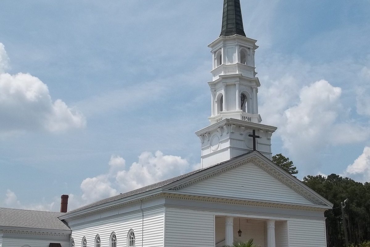 The white wooden Martha-Mary Chapel in Richmond Hill with its tall steeple, a cross on the gable and pine trees beside it under a cloudy sky