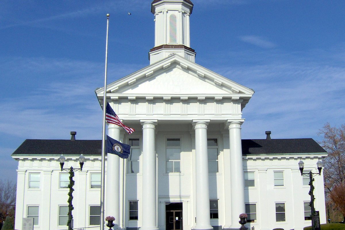 The white columned Madison County courthouse in Richmond with its cupola, an American flag and a Kentucky flag, under a blue sky