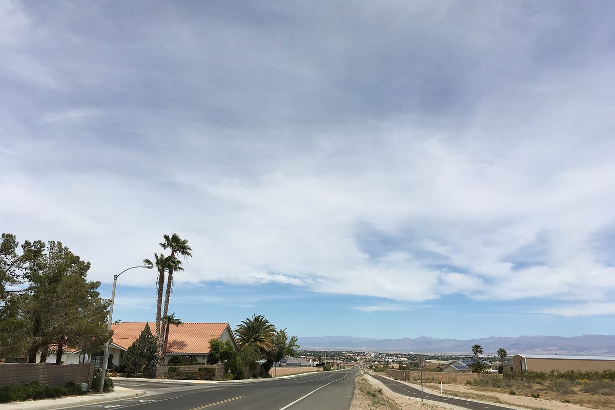 College Heights Boulevard in Ridgecrest running downhill past palm trees and a tile-roofed house toward desert mountains under a cloudy sky