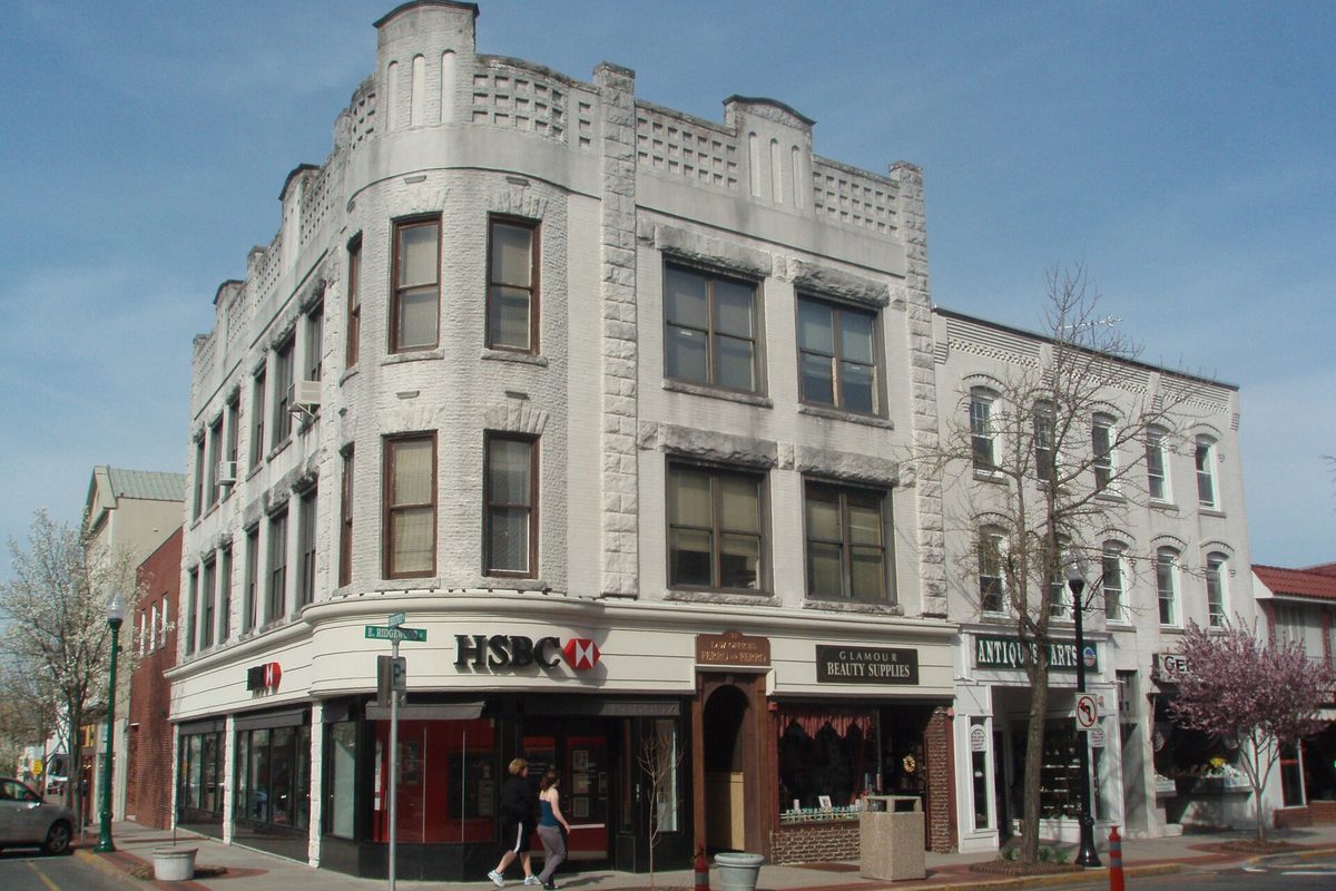 A pale stone three-story corner building with shops on the ground floor in downtown Ridgewood, with flowering trees and two people walking past