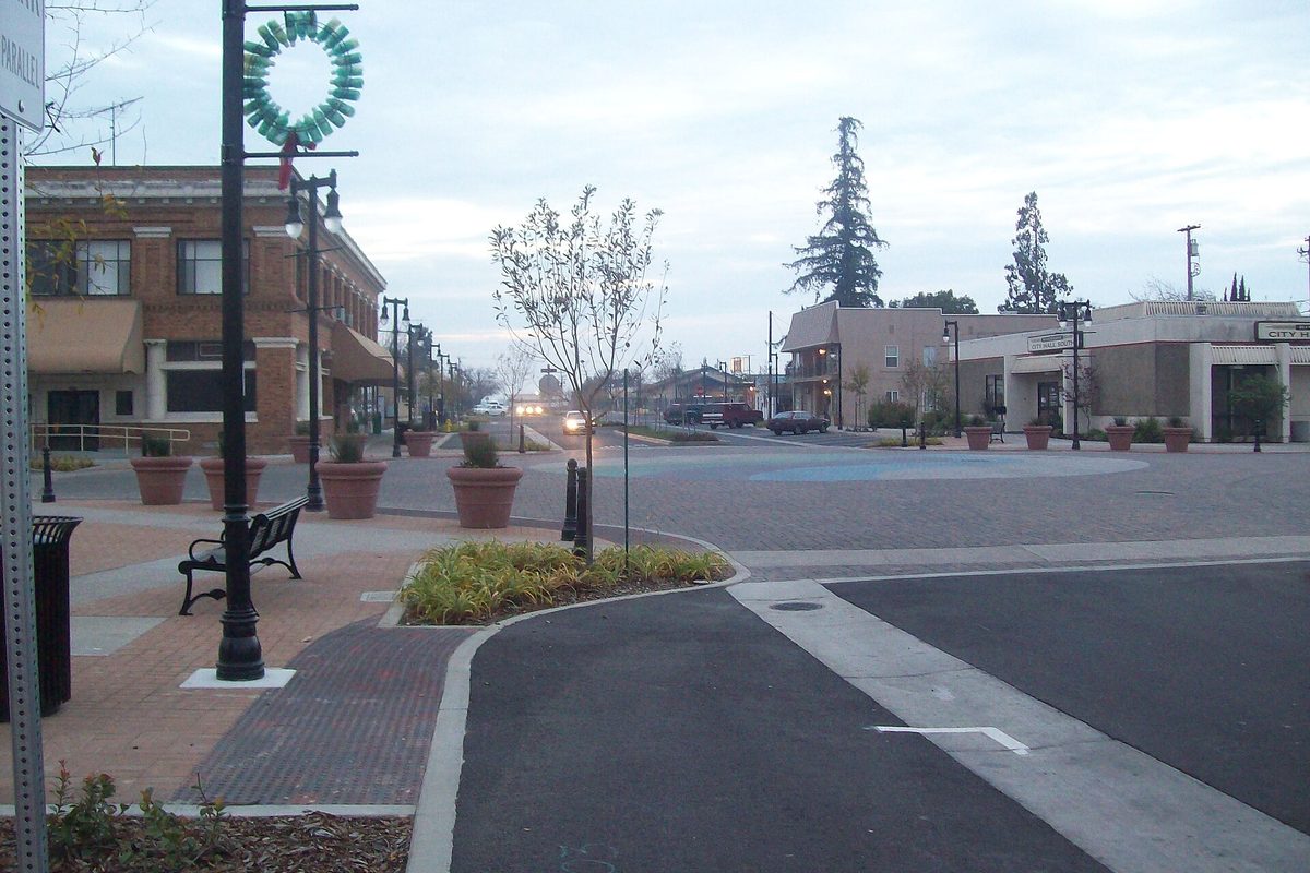 Third Street in downtown Riverbank at dusk, with a brick corner building, planters, a bench and street lamps hung with a holiday wreath