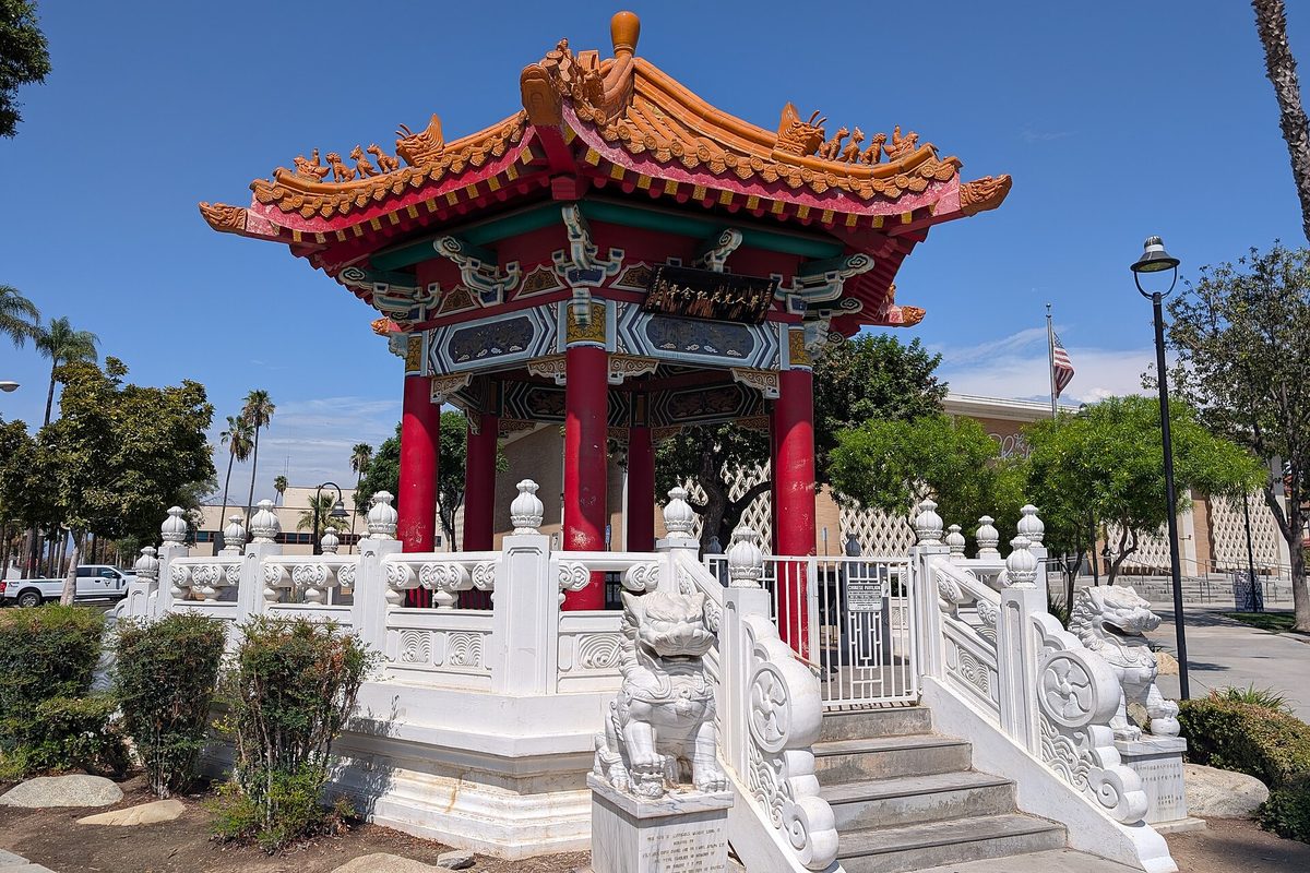 A red and gold Chinese pavilion with a curved tiled roof, ringed by white stone railings and guardian lion statues, among palm trees under a blue sky in Riverside