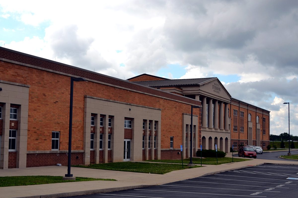 The long brick front of Stebbins High School in Riverside, with a columned entrance, a few cars parked beside the building, empty spaces in front and a cloudy sky