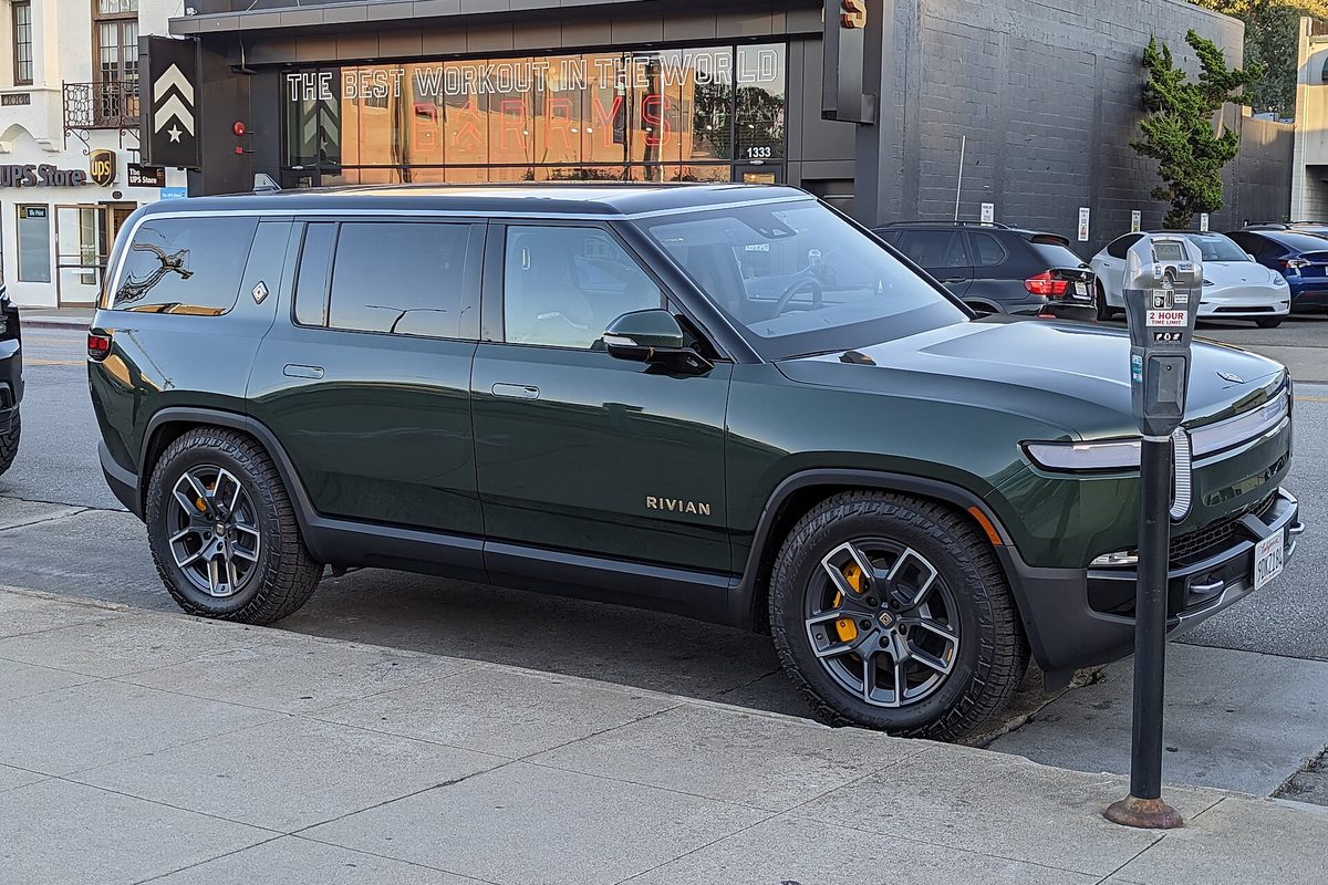 A dark green Rivian R1S SUV with yellow brake calipers, parked at a curb beside a parking meter on a city street