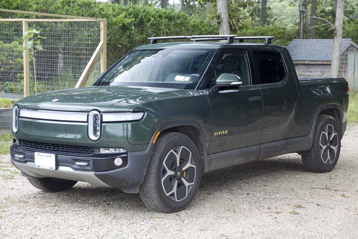 A forest green Rivian R1T pickup with a roof rack, parked on gravel in front of a fenced garden