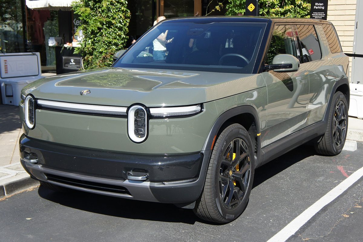 A sage green Rivian R2 SUV with black wheels and yellow calipers, parked in a marked space outside a shop
