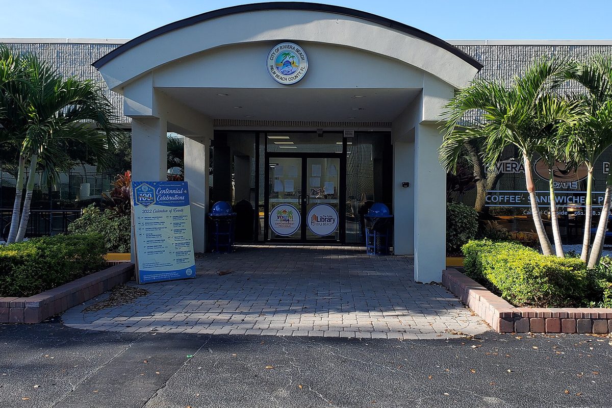 The arched entrance of the Riviera Beach Public Library, framed by palm trees, with a centennial celebration sign beside the door