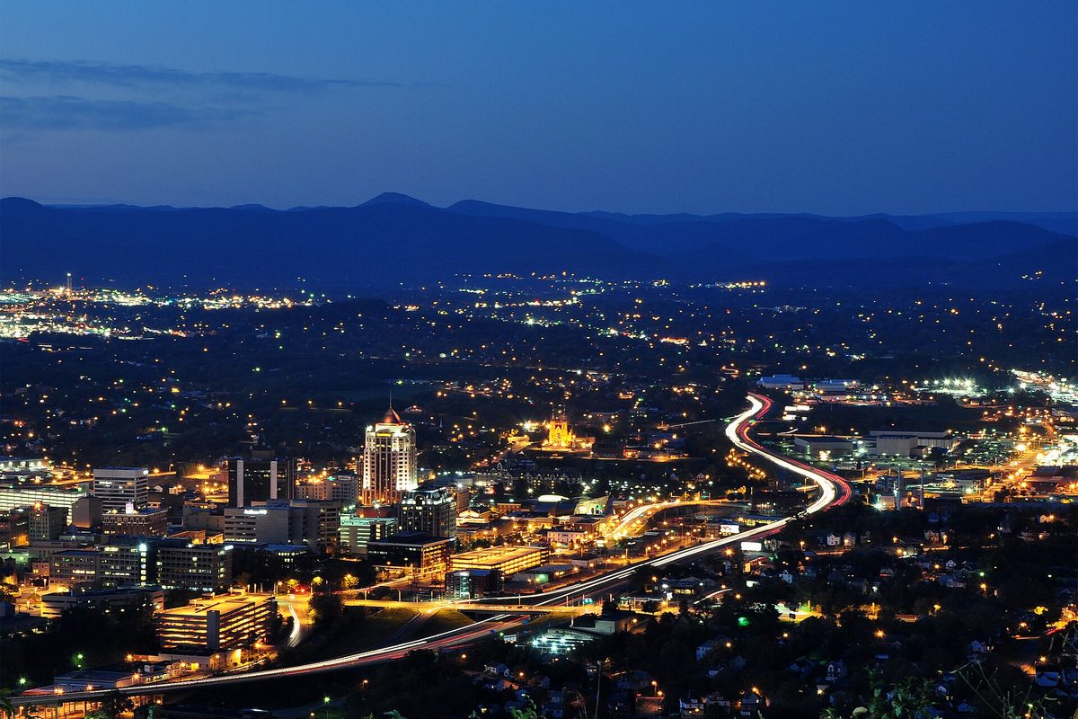 Roanoke at dusk seen from Mill Mountain: lit downtown towers, a highway drawn in streaks of headlights, and the dark ridges of the mountains behind