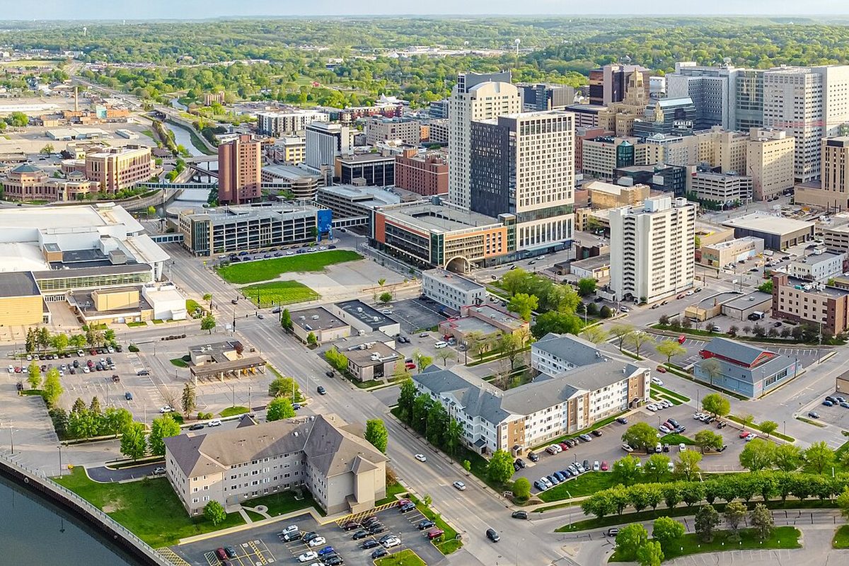 Aerial view of downtown Rochester, Minnesota, with tall office and hotel towers, apartment blocks, parking lots and leafy streets stretching toward green hills