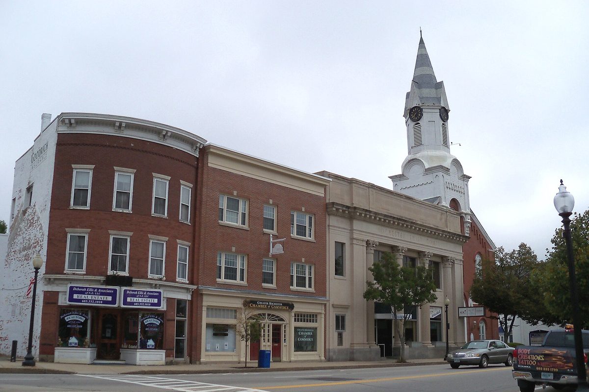 Downtown Rochester storefronts in red brick and pale stone, with a white church steeple and clock rising behind them on an overcast day