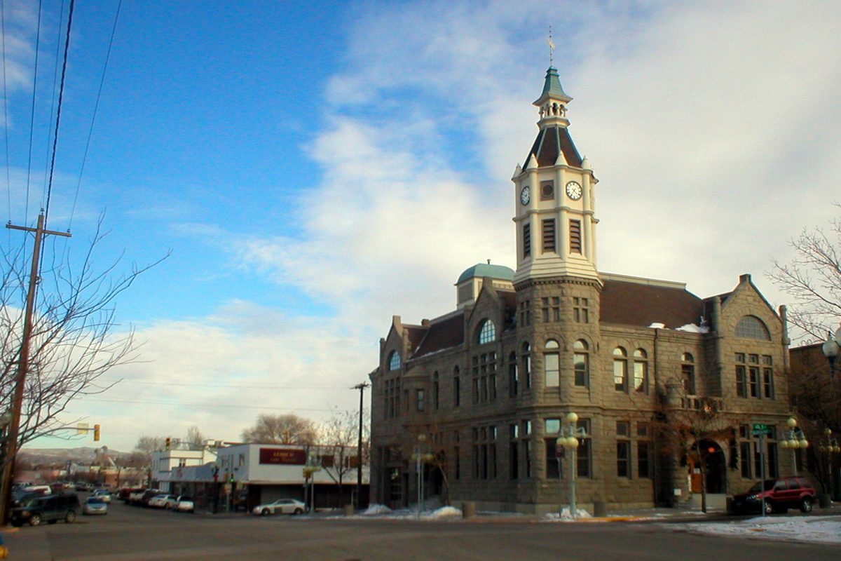 A sandstone building with a clock tower and green cupola on a corner in historic downtown Rock Springs, with parked cars and patches of snow