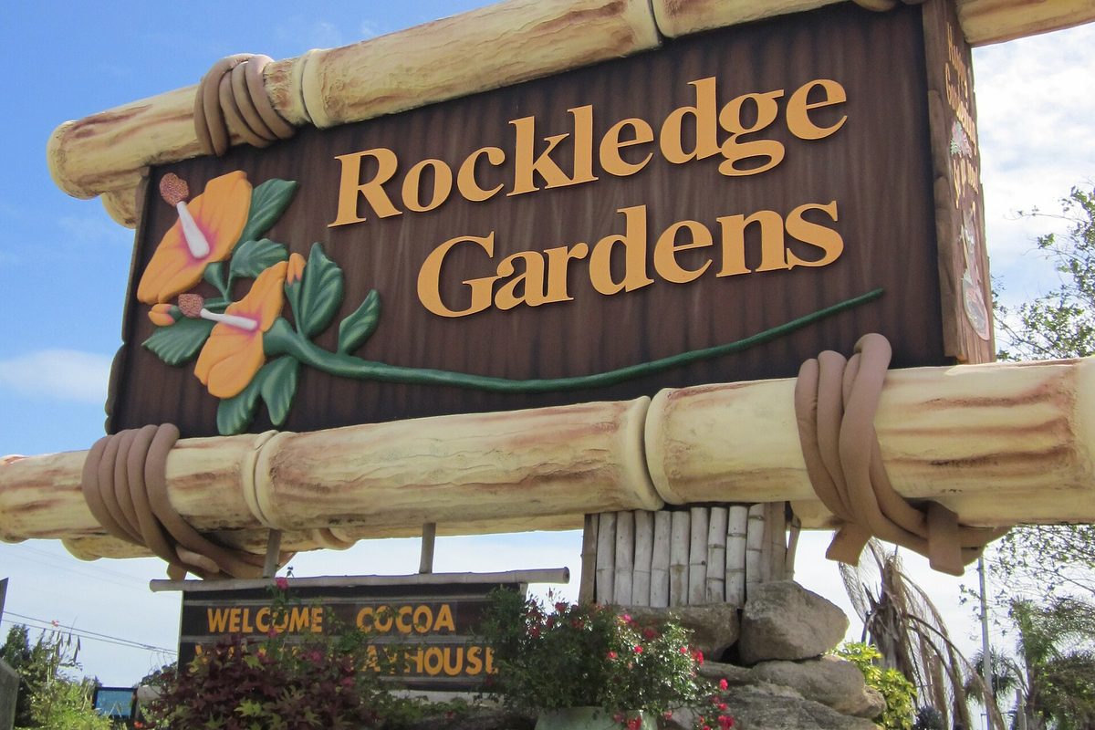 A carved wooden Rockledge Gardens sign painted with orange hibiscus flowers, framed by faux bamboo poles above flowering shrubs