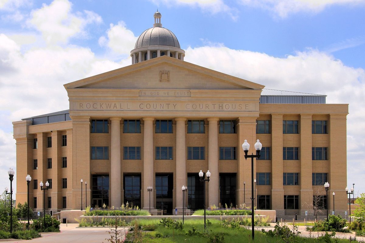 The Rockwall County Courthouse, a tan stone building with tall columns and a silver dome, behind planted beds and black lamp posts