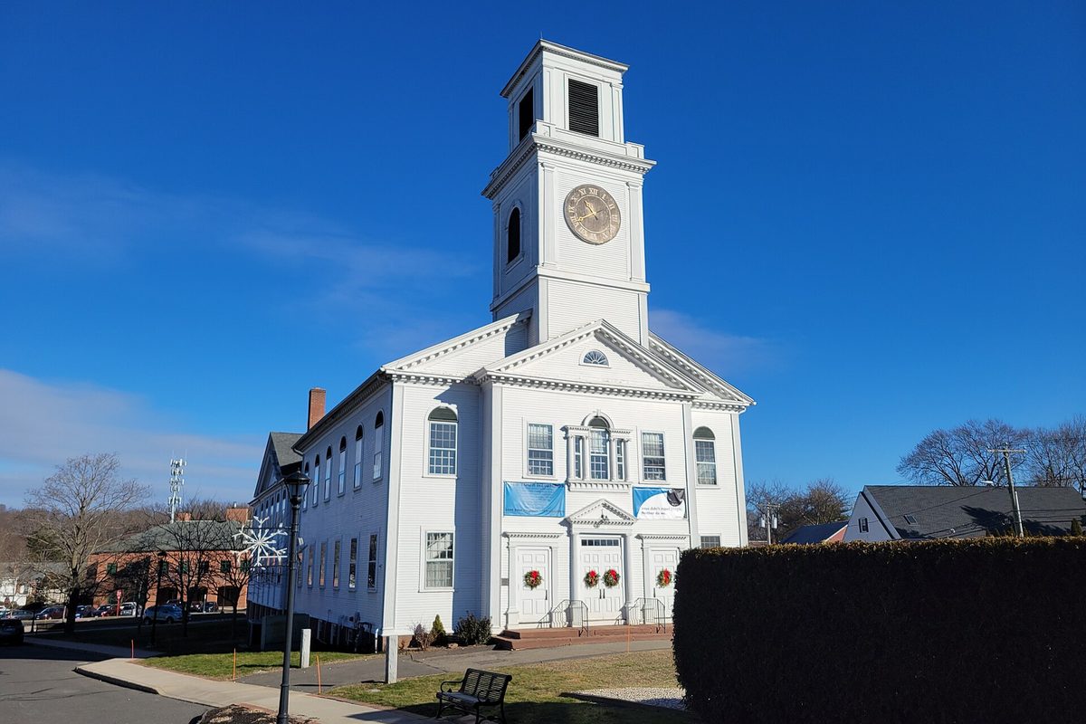 The white Rocky Hill Congregational Church with a tall clock tower, wreaths on its doors and a bench and lamppost out front, under a deep blue sky
