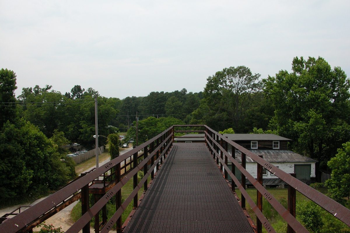 A rust-brown steel pedestrian bridge with a metal grate walkway crossing the railroad in Rocky Mount, with green trees and a white frame building below