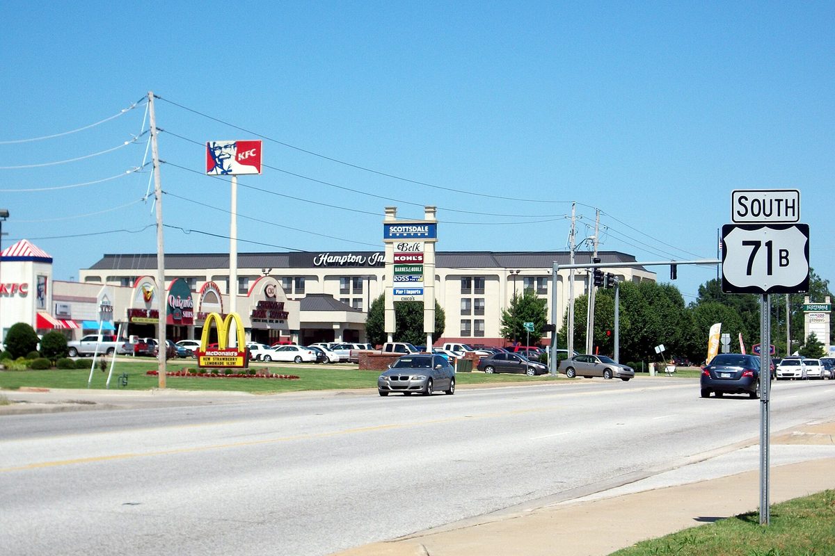 A US 71B South sign beside a wide road in Rogers, with a Hampton Inn, a KFC and a McDonald's along the roadside under a blue sky