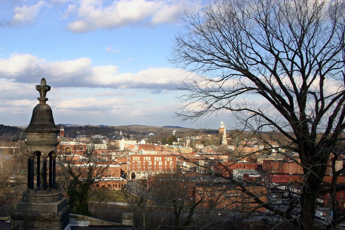 Downtown Rome's brick buildings and church towers seen from Myrtle Hill Cemetery, with a bare tree and a stone monument in the foreground and hills on the horizon