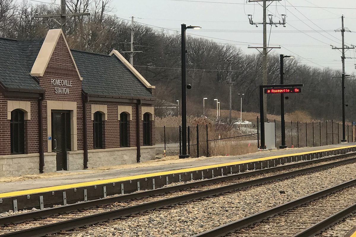 The brick Romeoville Metra station beside the tracks, with a red Romeoville sign over the platform and bare trees behind on an overcast day