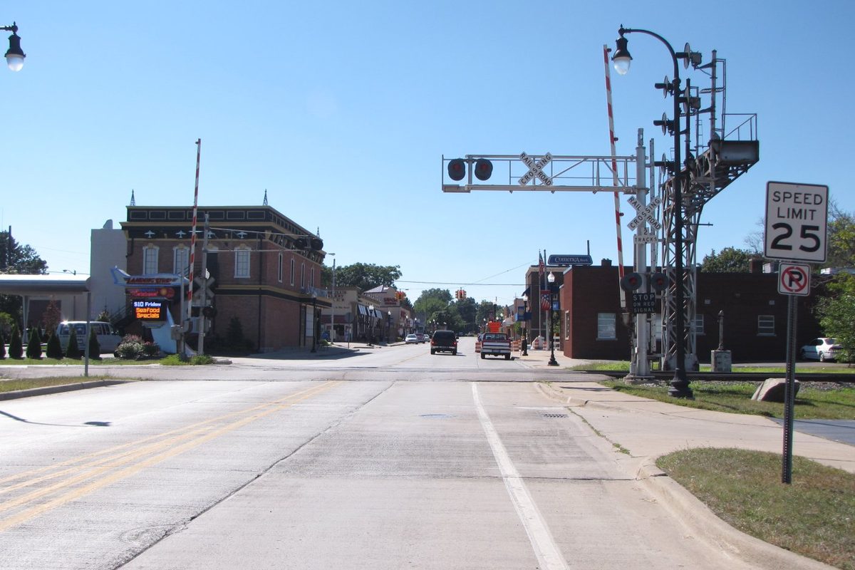 Downtown Romulus street with railroad crossing signals and gates, brick storefronts on both sides and a 25 mph speed limit sign under a clear blue sky