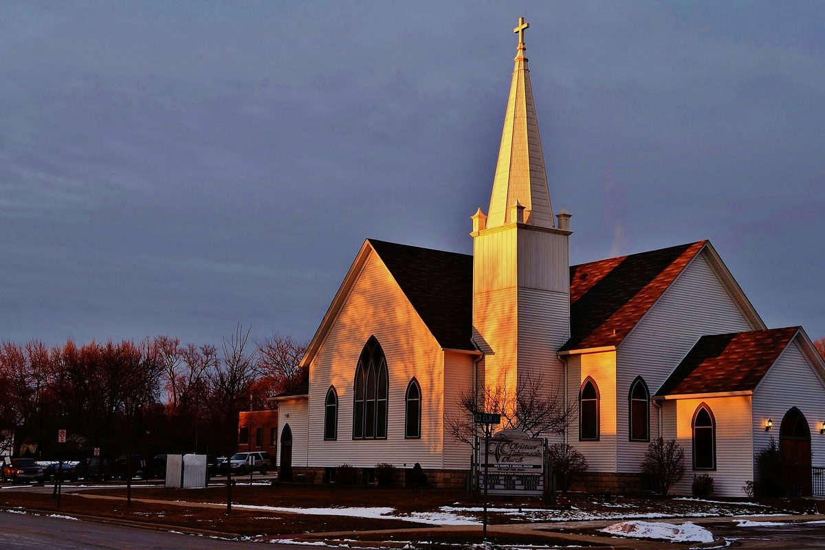 The white wooden Christian Tabernacle Church in Roselle, with a tall steeple and pointed arched windows, lit orange by low sun under a gray sky, with patches of snow on the lawn