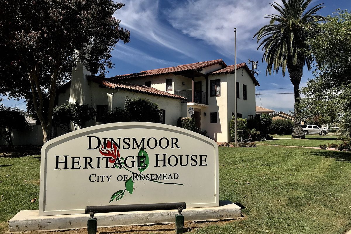 A sign reading Dinsmoor Heritage House, City of Rosemead, with a painted rose, in front of a white two-story house with a red tile roof and a tall palm