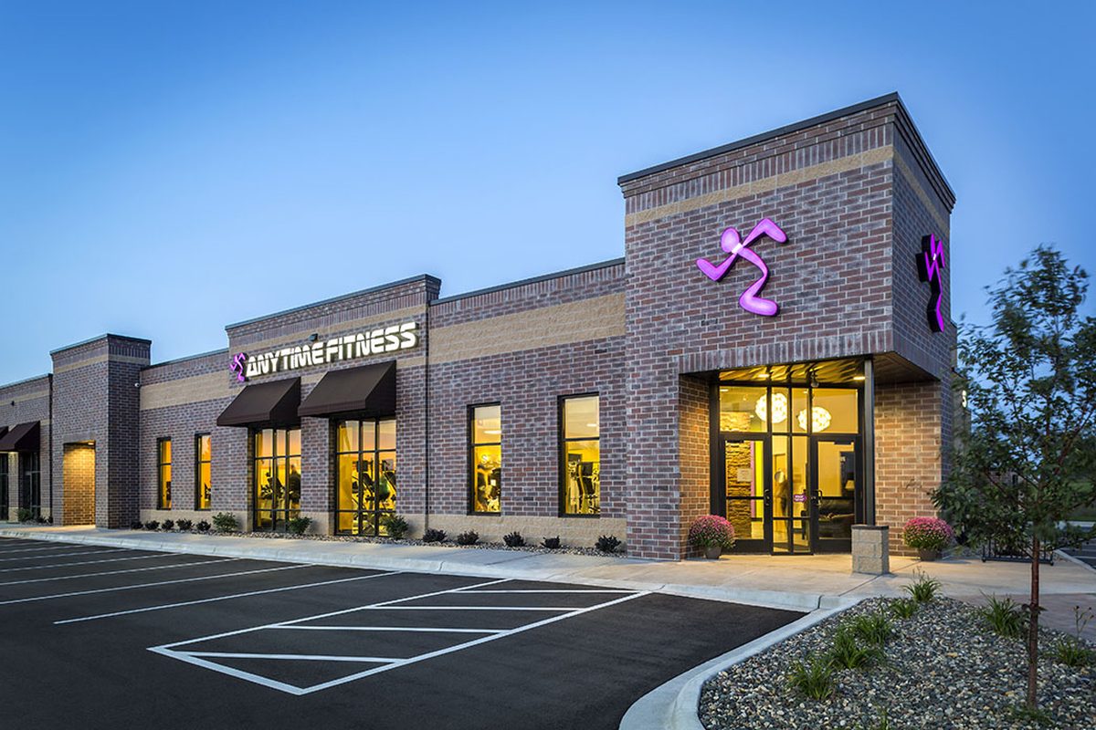 A one-story brick fitness center in Rosemount at dusk, its windows lit, with purple signs, black awnings and an empty parking lot in front