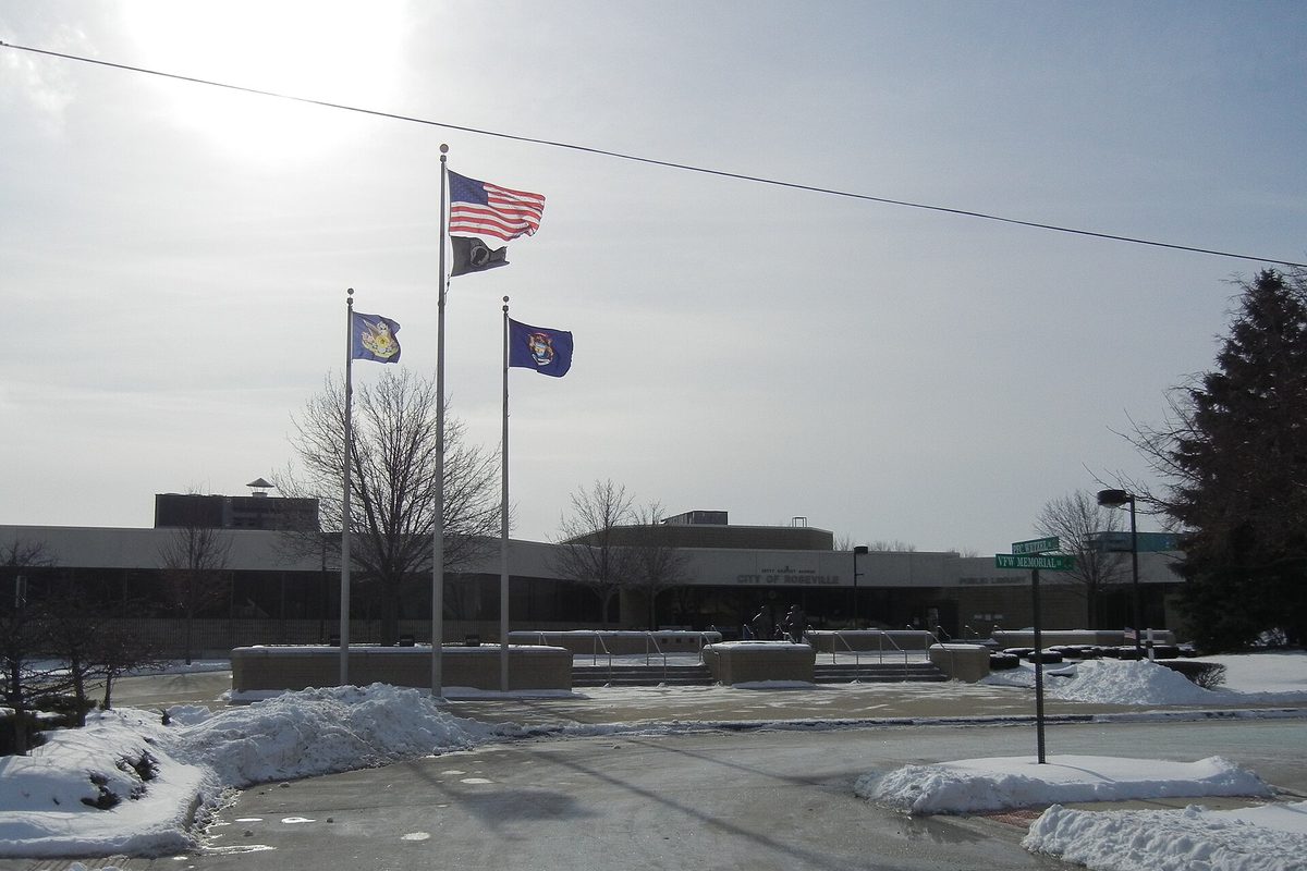 Flags on tall poles in front of the long, low Roseville Civic Center on a winter day, with snow piled along the drive
