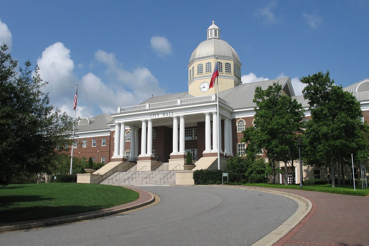 Roswell City Hall, a white-columned building with a domed clock tower and an American flag, at the end of a curved drive under a blue sky