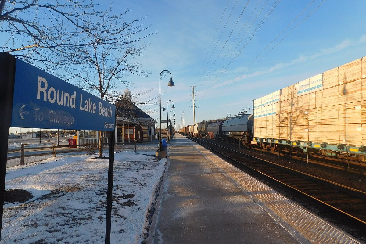 The blue Round Lake Beach station sign on a snowy Metra platform, with the station building behind it and a freight train passing on the tracks