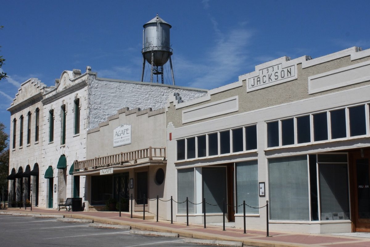 A row of old white stone and stucco storefronts in downtown Round Rock, one with Jackson on its cornice, with a metal water tower against a blue sky behind them