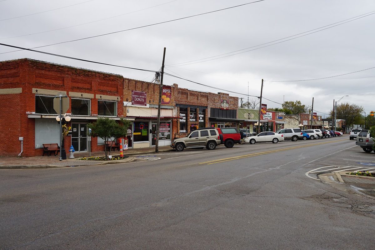 Main Street in Royse City lined with old red-brick storefronts, a donut shop sign and parked pickups on an overcast day