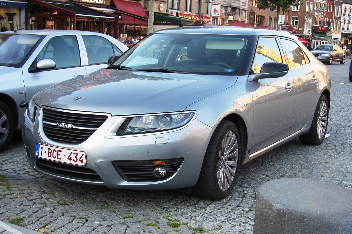 A silver second-generation Saab 9-5 sedan parked on a cobblestone square in front of shops and cafes