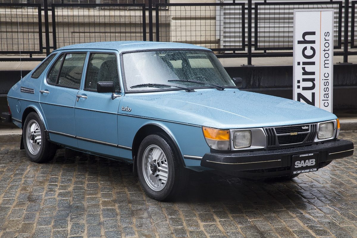 A pale blue classic Saab 900 Turbo five-door parked on wet cobblestones outside a classic car dealer