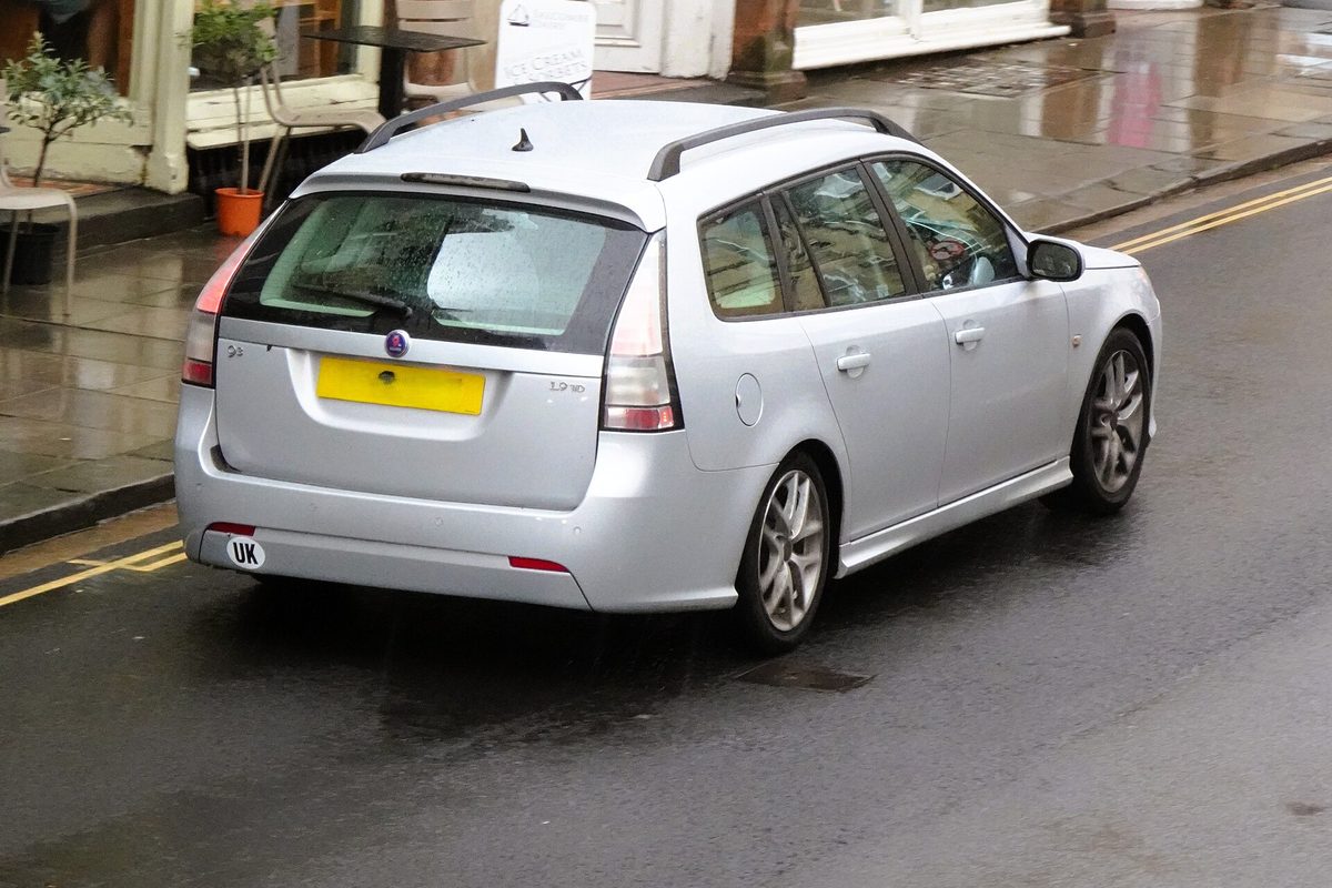 A silver 2007 Saab 9-3 estate seen from behind and to one side, driving down a wet town street past a shopfront