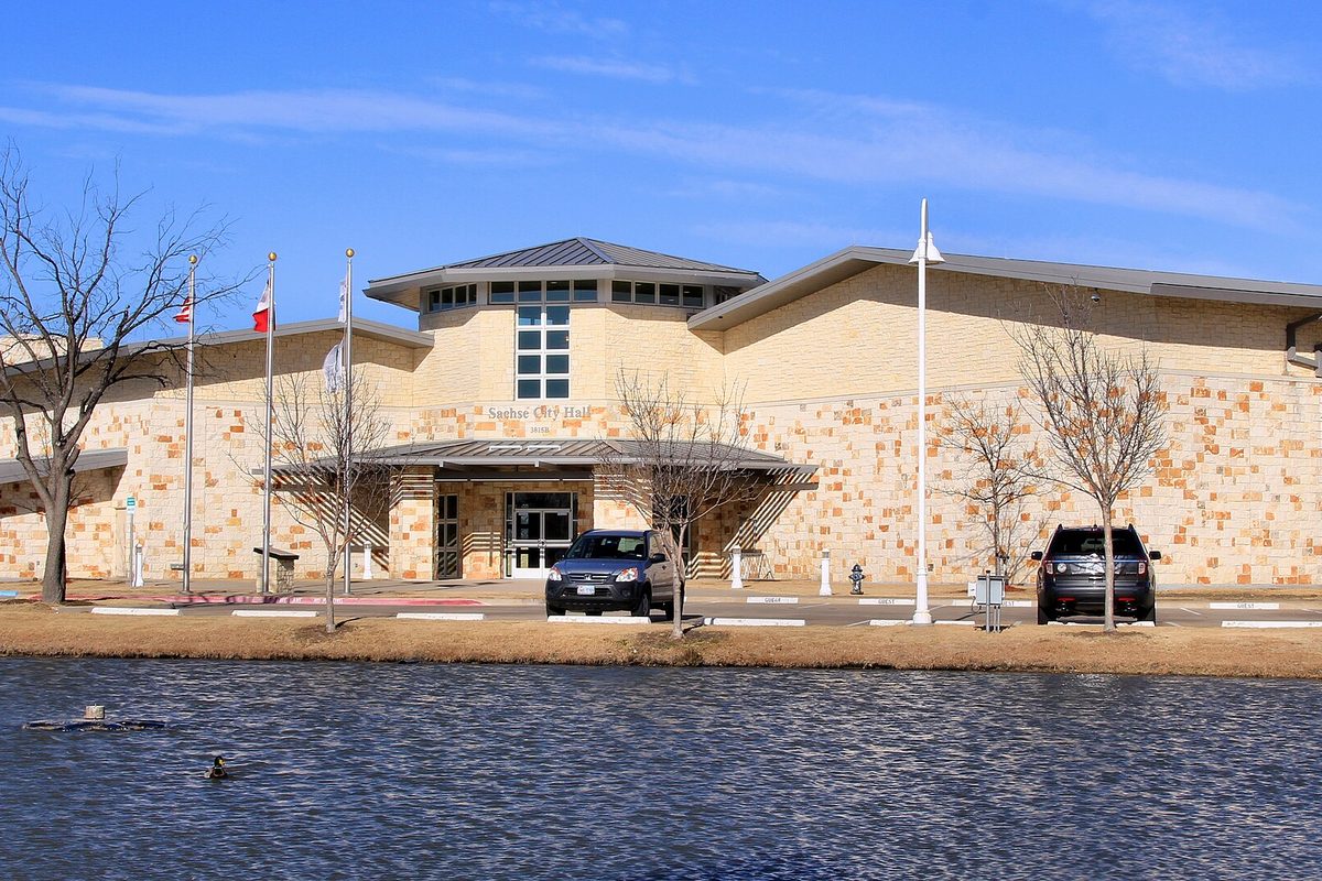 Sachse City Hall, a long tan stone building with flagpoles and two parked SUVs, seen across a pond on a clear winter day