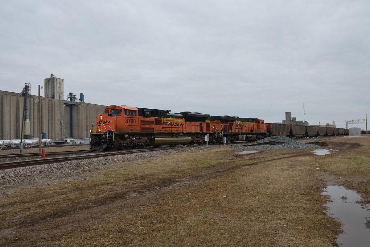 An orange BNSF locomotive leading a long coal train past tall grain elevators in Saginaw under a gray sky