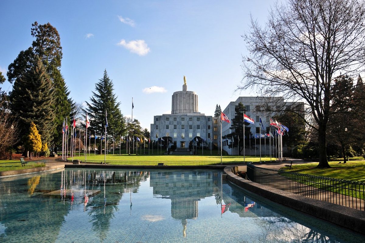 The Oregon State Capitol with its gold statue on top, reflected in the Waite Fountain pool and framed by flags and evergreens