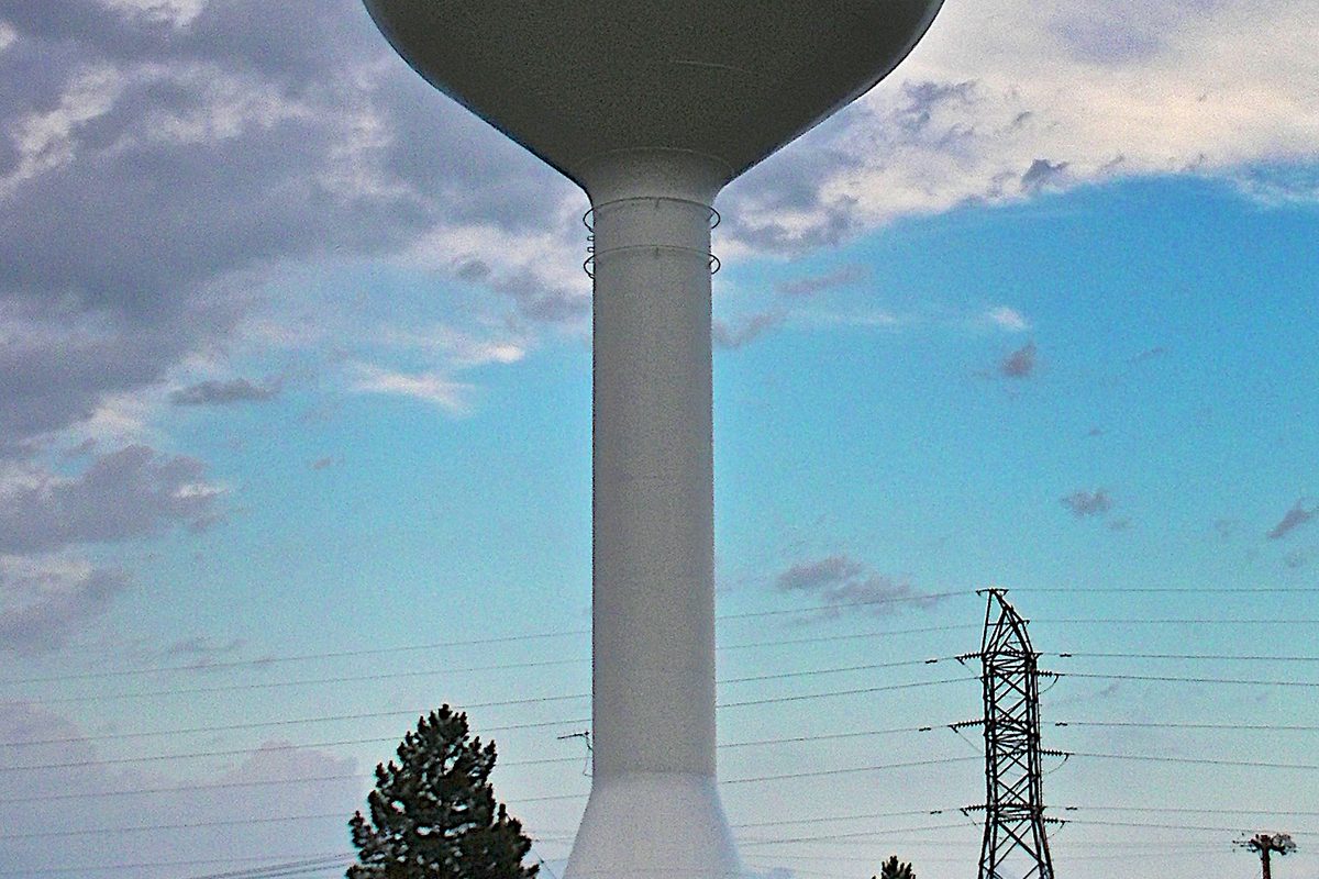 A white water tower on Salina's southeast side against a blue sky with scattered clouds, with a pine tree and a power line tower beside it