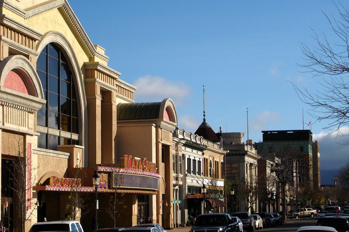 A downtown Salinas street with an arched cream-colored theater building, older storefronts and parked cars under a blue sky