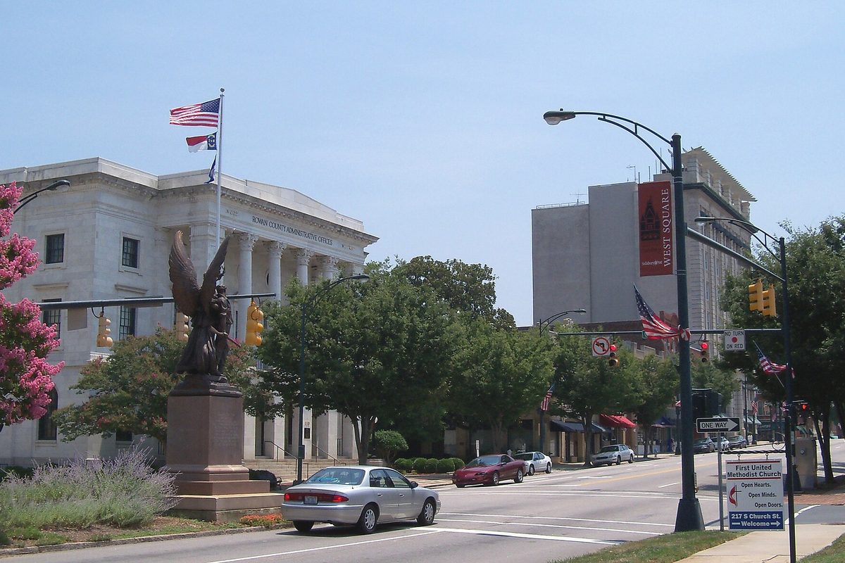 A downtown Salisbury street with a bronze winged statue on a stone pedestal, a white columned building flying flags, and cars at a traffic light