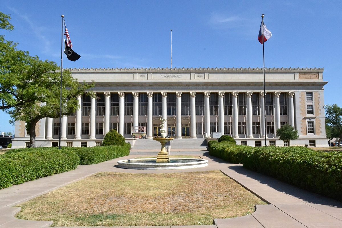 The white columned front of the Tom Green County Courthouse in San Angelo, with a fountain, trimmed hedges and three flags in front under a blue sky